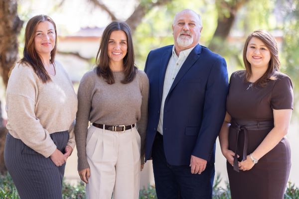 Admin team photo. Four people standing outdoors in front of trees, dressed in business-casual and professional attire, smiling at the camera in natural daylight.