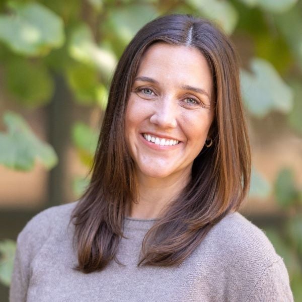 A woman with long brown hair stands outdoors in front of a blurred background of leafy vines. She is smiling at the camera and wearing a light-brown sweater tucked into cream-colored pants with a brown belt.