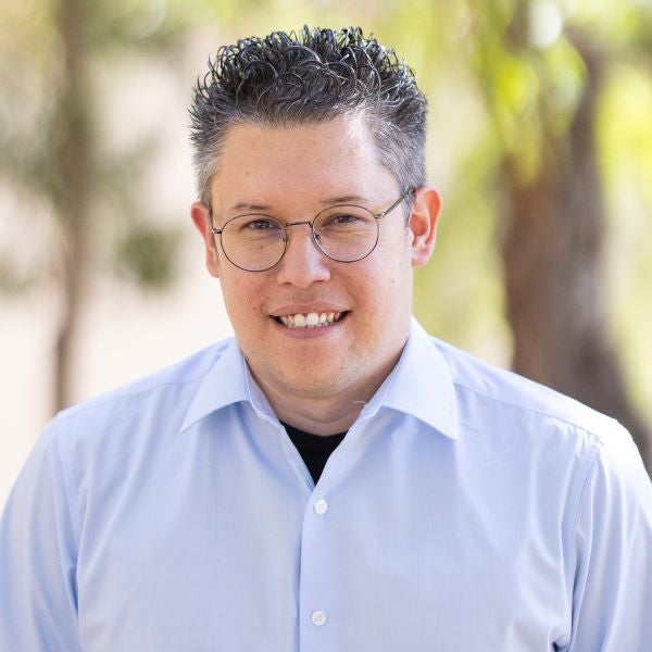 Erik Noriega. Smiling person with short curly hair and glasses wearing a light blue button-down shirt, standing outdoors with softly blurred trees and sunlight in the background.
