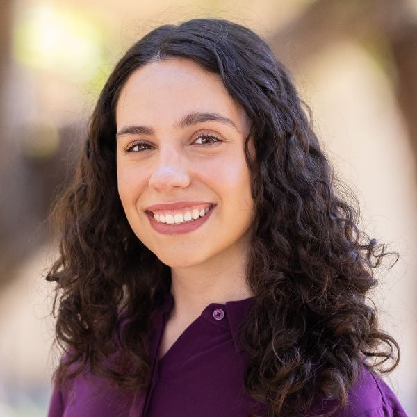 A woman with long curly dark hair stands outdoors, smiling at the camera. She is wearing a long-sleeved plum-colored blouse tucked into black pants with a brown braided belt. Behind her are green shrubs and the soft-focus trunks of large trees, with warm sunlight creating a natural, inviting background.