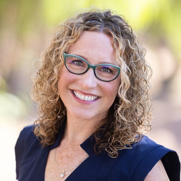 Lauren Bianchi Klemann. Portrait of a smiling woman with shoulder-length curly blonde hair, wearing green-rimmed glasses and a navy blue top, standing outdoors with a softly blurred natural background.