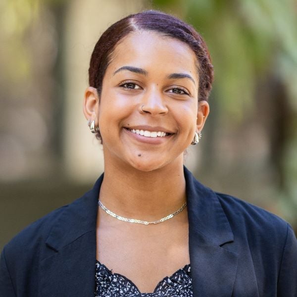headshot of Mia Lewis. Smiling woman wearing a black blazer over a patterned top, accessorized with hoop earrings and a delicate chain necklace, standing outdoors with a softly blurred background of greenery and light.