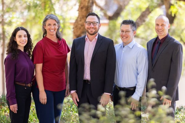 Multimodal Programs Division team. Five people standing outdoors in front of leafy trees, dressed in business and business-casual attire, smiling at the camera in natural daylight.