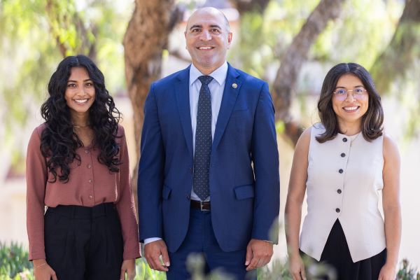 Programming Division. Three people standing outdoors in front of trees, dressed in business and business-casual attire, smiling at the camera in natural daylight.