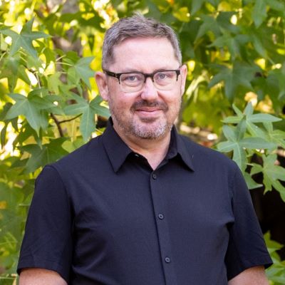Andrew Orfila with short hair and glasses wearing a black collared shirt, standing outdoors in front of green leafy trees, smiling at the camera.
