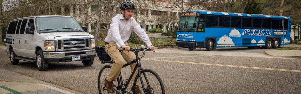 Andrew wearing a helmet and white shirt rides a bicycle on a street, with a white commuter van and a blue 'Clean Air Express' bus bound for Santa Maria visible in the background.