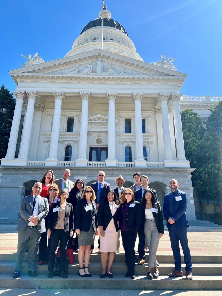 Sarkes Khachek stands at the far right of a group of colleagues on the front steps of the California State Capitol in Sacramento. The group, dressed in professional business attire, smiles in the bright sunlight with the historic white Capitol building and its grand columns rising behind them.