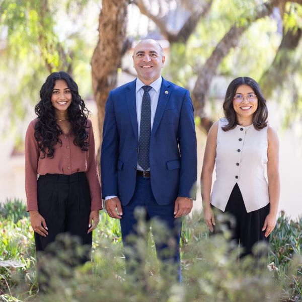 Sarkes Khachek stands outdoors with two members of the SBCAG programming team, smiling in front of leafy trees and soft greenery. Sarkes, wearing a blue suit and patterned tie, is centered between a team member in a rose-brown blouse and black pants on the left and another in a sleeveless cream top and black skirt on the right.