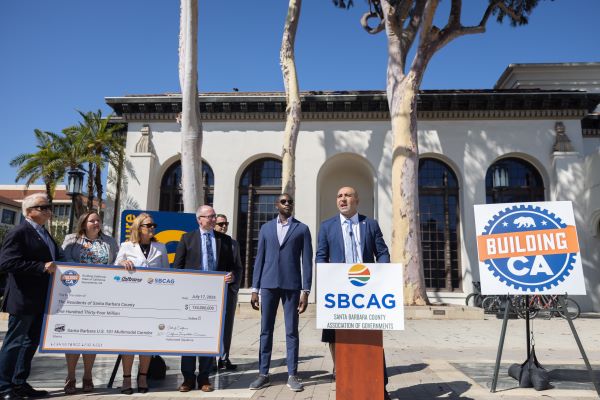 Sarkes Khachek speaks at an outdoor press event in front of a historic Santa Barbara building, standing at a podium with the SBCAG logo. To his left, a group of officials and community leaders hold an oversized ceremonial check displaying $134 million in SB1 funding for the U.S. 101 Multimodal Corridor. A large “Building CA” sign on an easel stands to the right, and tall palm trees frame the sunny scene.
