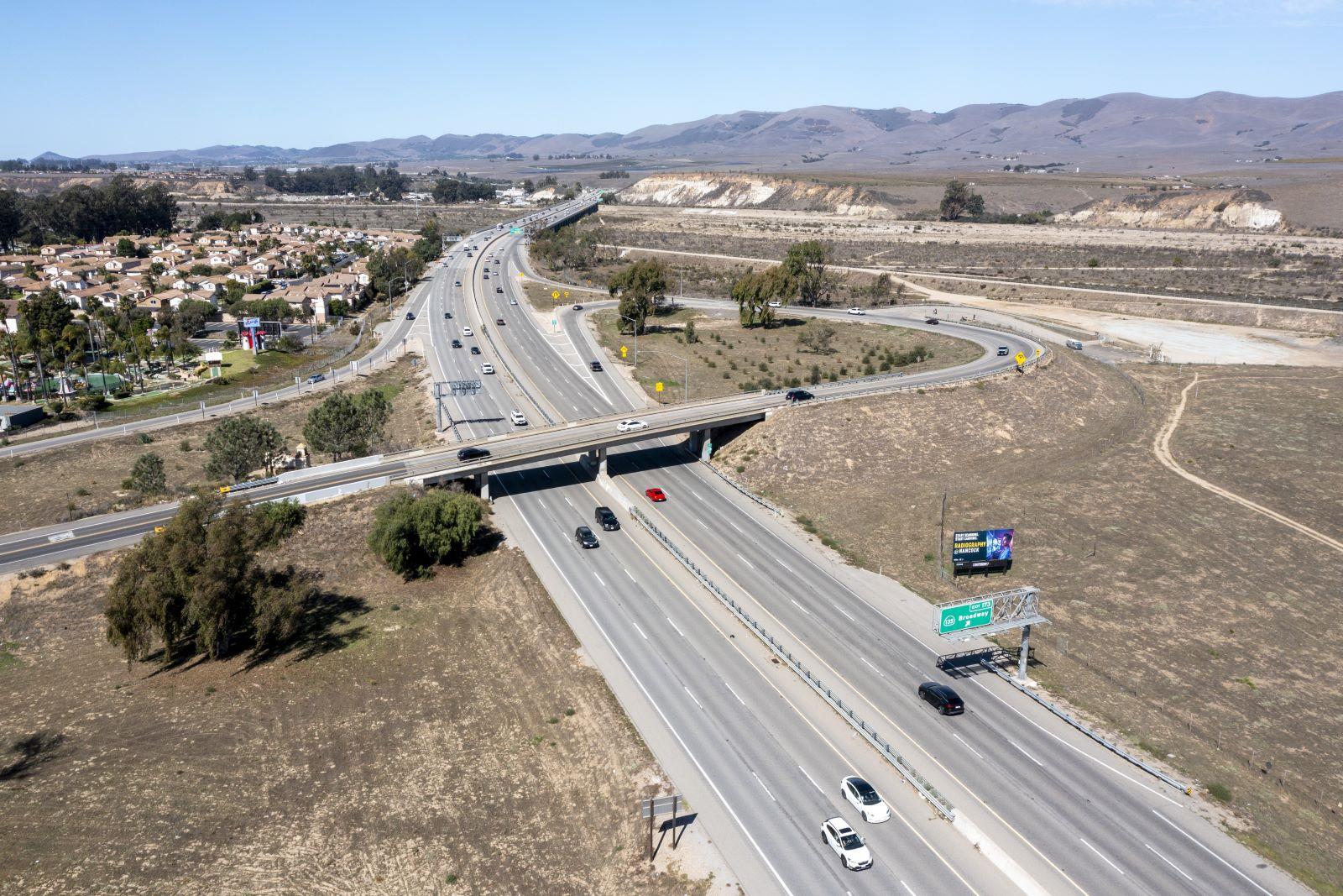 Aerial view of the U.S. Highway 101 corridor in Santa Maria, showing the State Route 135 overcrossing and interchange area with multiple freeway lanes, vehicles traveling in both directions, and surrounding open fields, hills, and nearby residential neighborhoods.