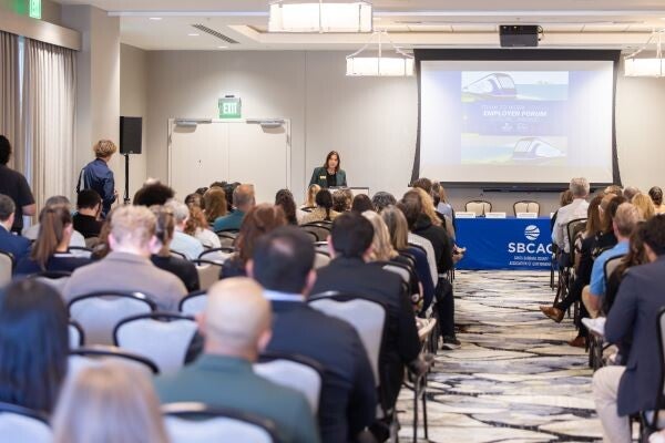 Audience members sit in rows during a public forum as a speaker presents at a podium, with an SBCAG information table nearby.